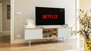 Un salon moderne avec un téléviseur à écran plat affichant le logo « NETFLIX », un meuble en bois en dessous et un vase avec des fleurs séchées sur la droite.