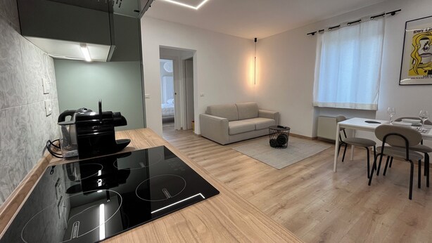 Una cocina moderna con una placa de inducción negra, un comedor con mesa y sillas blancas, y una sala de estar con un sofá y una mesita auxiliar.