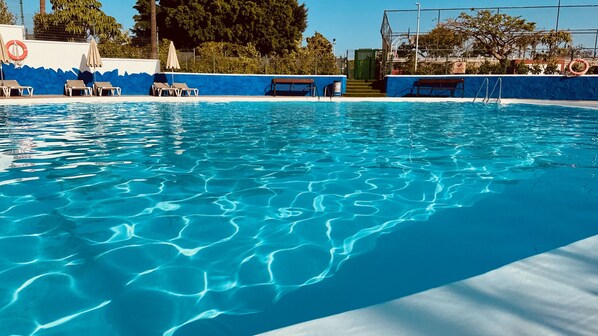 Een helder zwembad met blauw water, omringd door een blauwe muur en een hek. Er zijn ligbedden en een bank langs het zwembad.