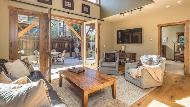A spacious living room with a wooden coffee table, leather sofa, and armchairs. Large windows allow natural light and provide views of the outdoor patio area. - Walk to Ski Shuttle and Town | Hot Tub (Big Sky)