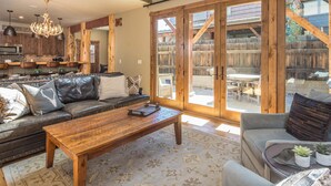 A spacious living area with a leather sofa, a wooden coffee table, and a grey armchair. There is a large sliding glass door leading to an outdoor patio with wooden furniture. - Walk to Ski Shuttle and Town | Hot Tub (Big Sky)