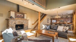 A spacious living room with a stone fireplace, a wooden staircase, and a modern kitchen with wooden cabinets. - Walk to Ski Shuttle and Town | Hot Tub (Big Sky)