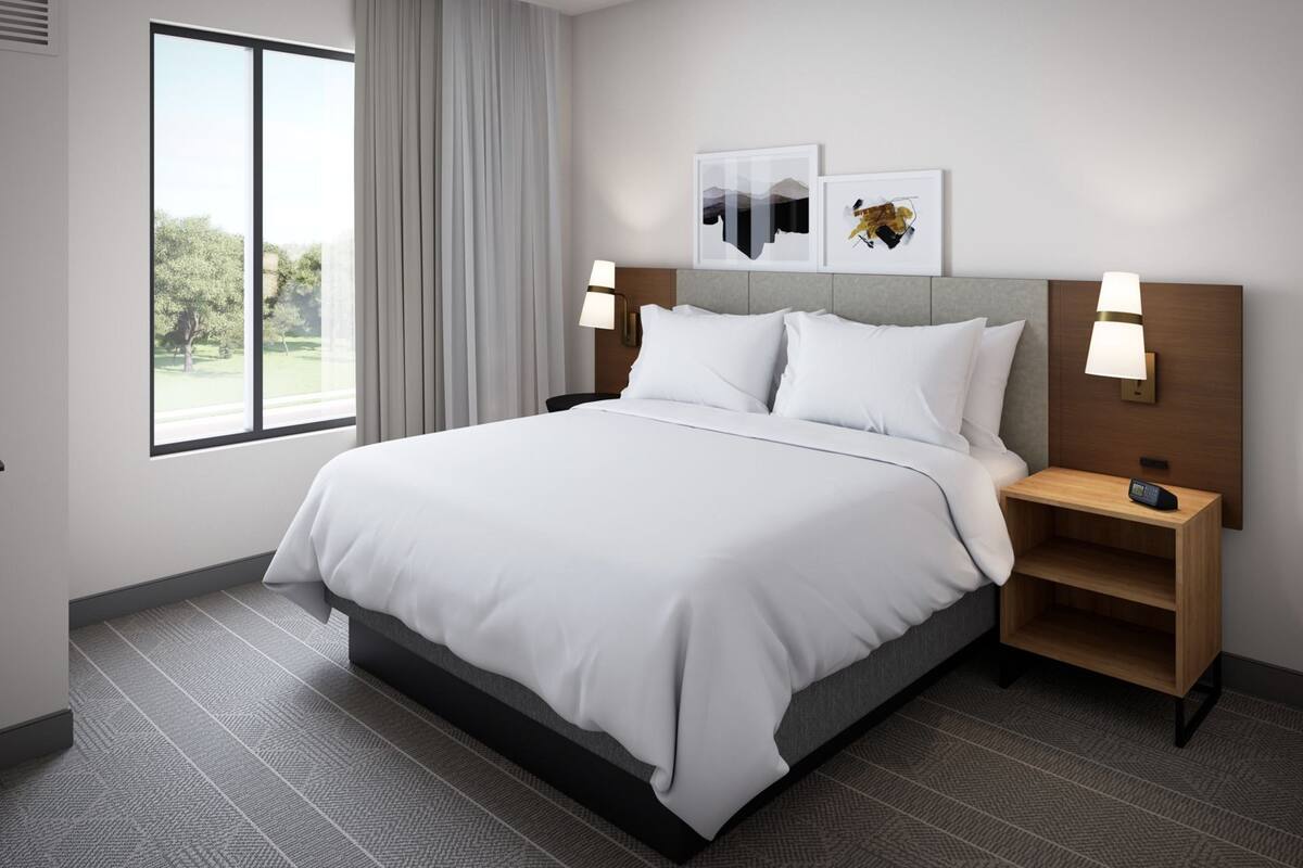 A hotel room with a large bed, two bedside tables, a window with curtains, and artwork on the wall.