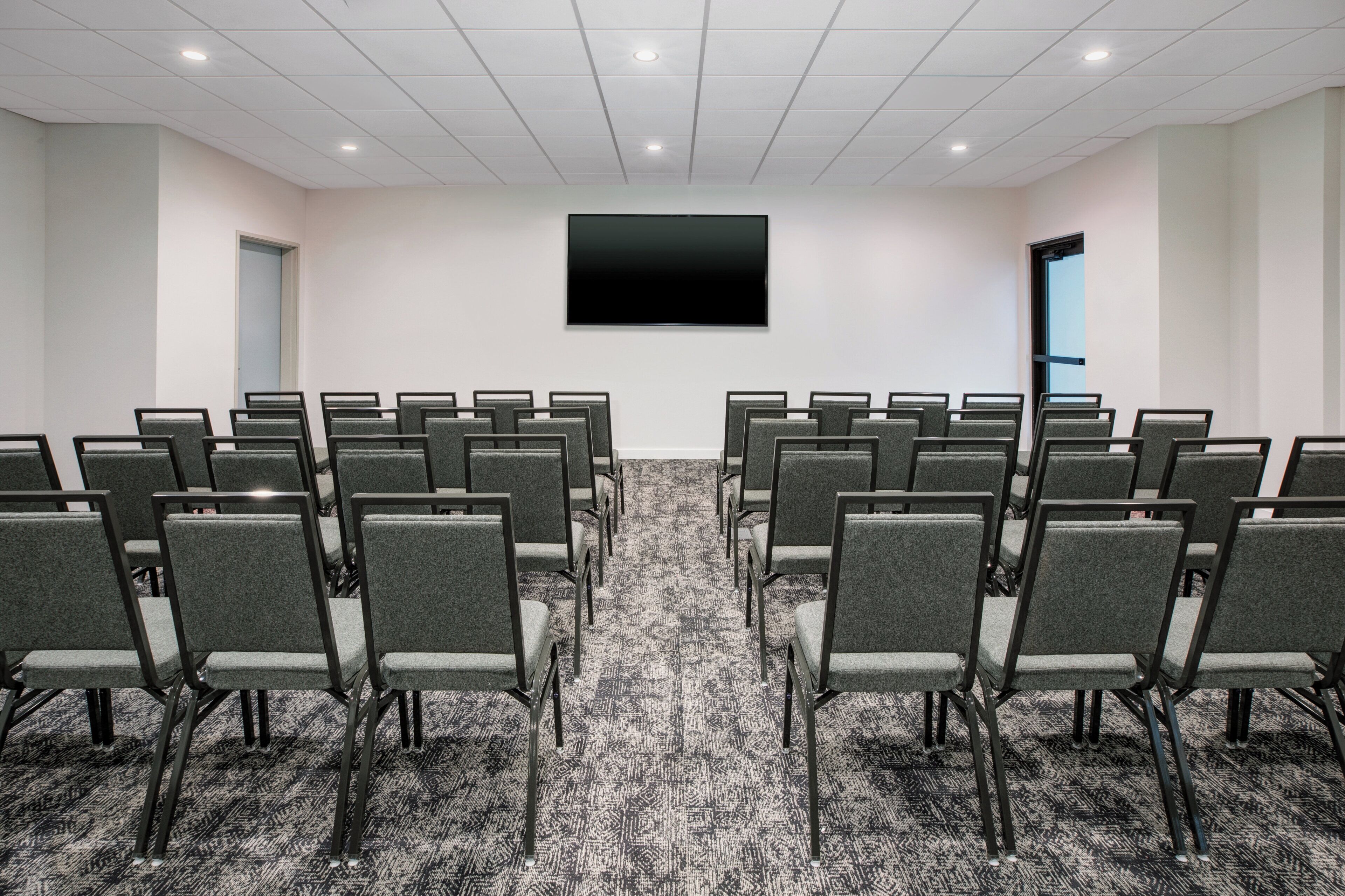 A conference room with rows of chairs, a large flat-screen TV on the wall, and carpeted flooring.