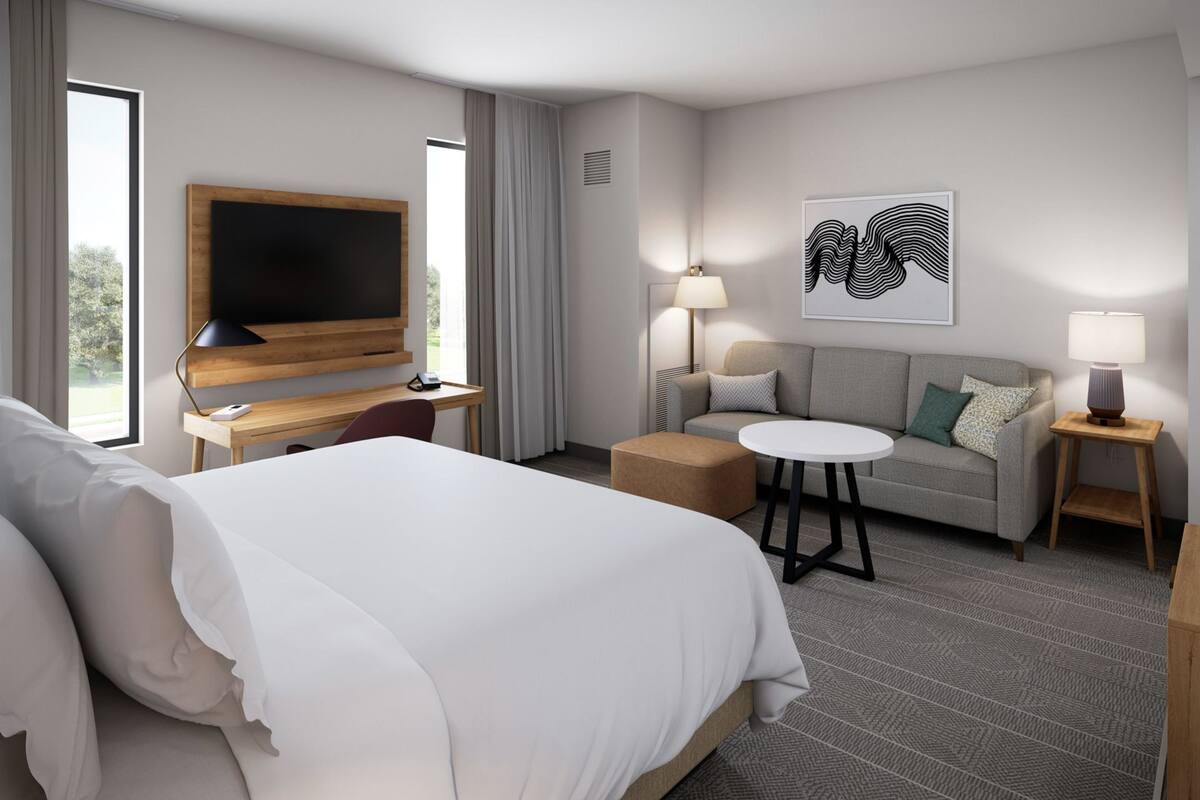 A hotel room with a large bed, a desk with a flat-screen TV, a sofa, a small round table, and a lamp.