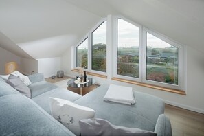 A modern living room with a high ceiling and large windows offering a view of greenery and buildings.