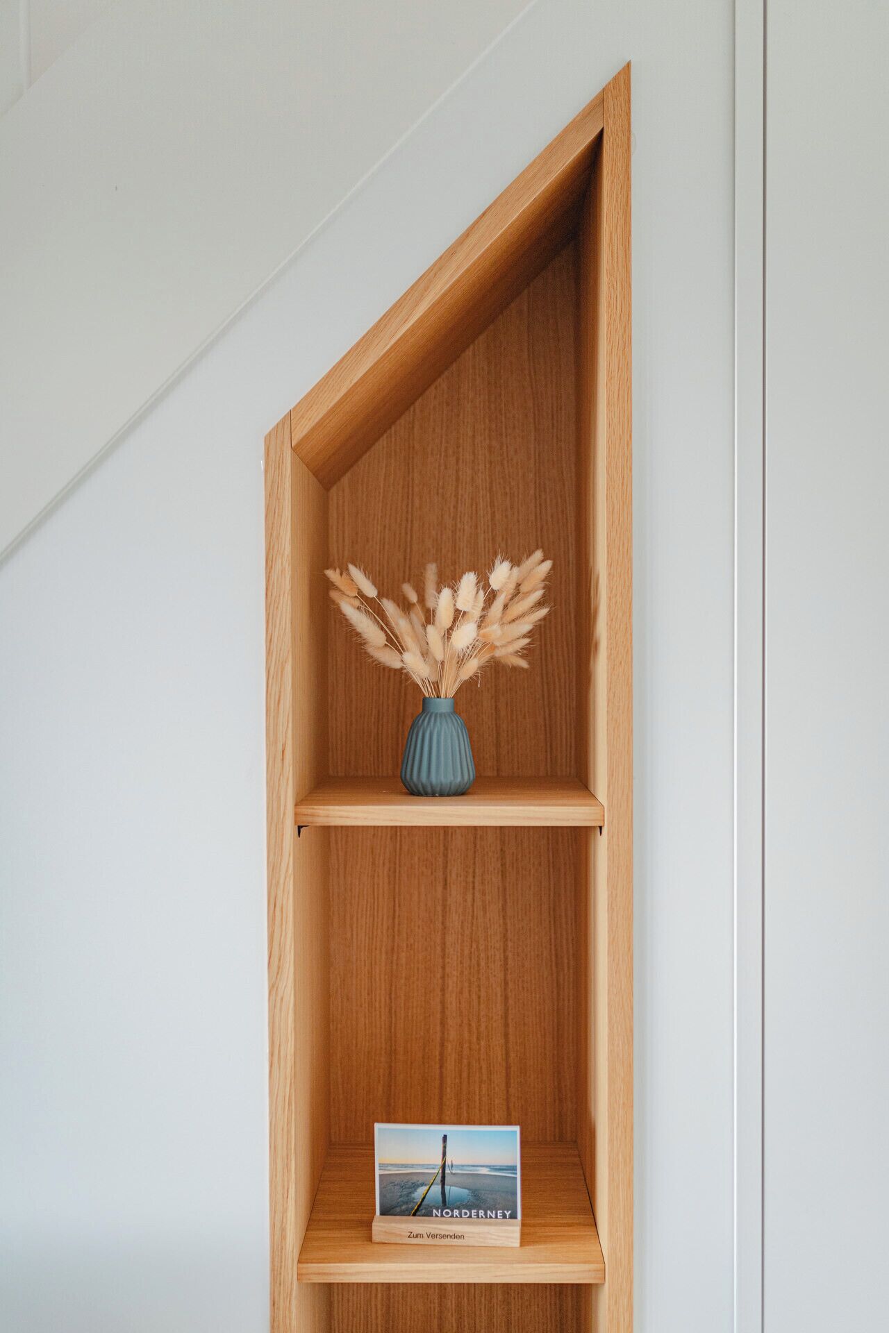 Une étagère en bois ornée d’un vase contenant des herbes de la pampa séchées et d’un livre intitulé « Norderney Zum Versenden ».