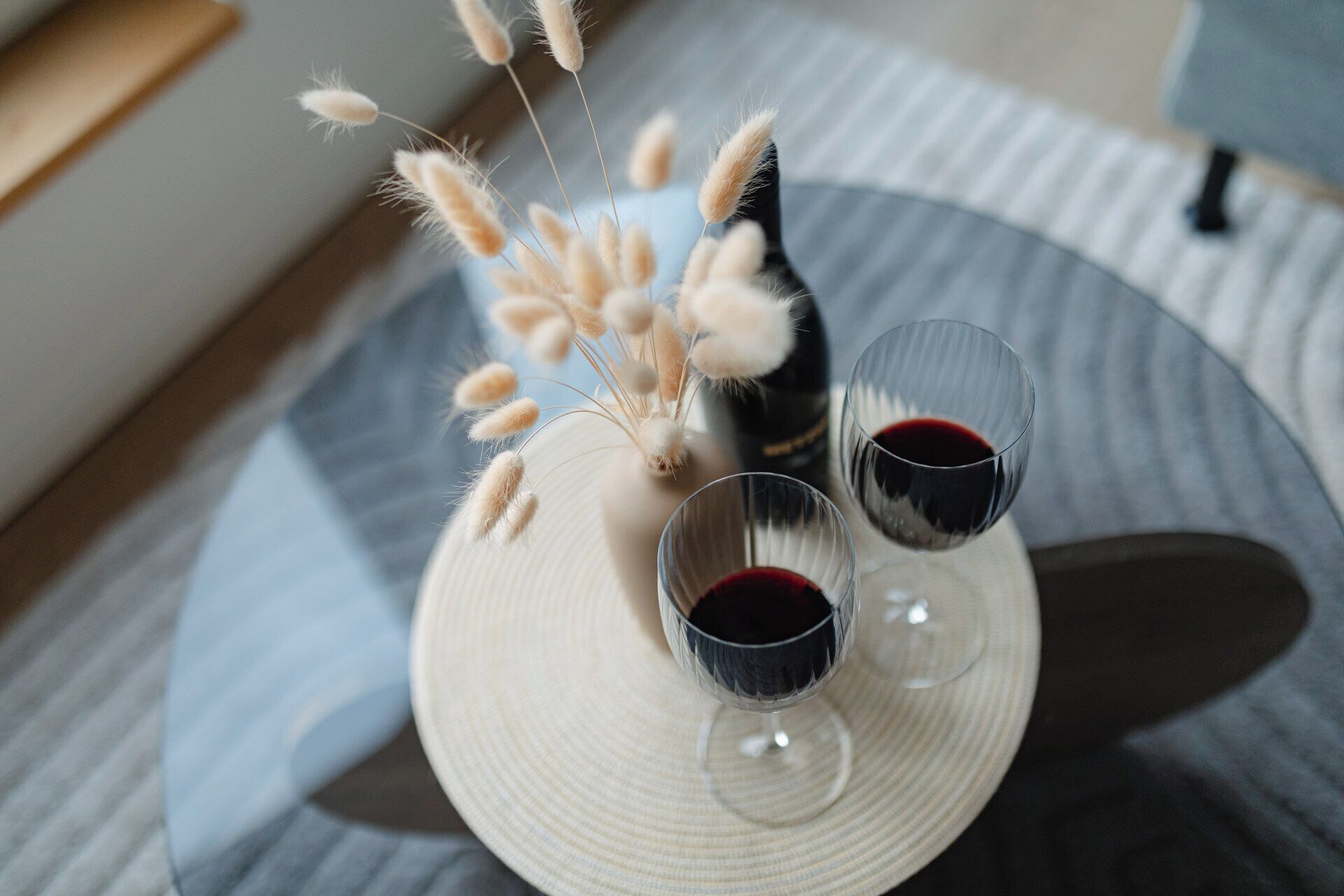 Une table ronde sur laquelle sont posés un vase blanc et deux verres de vin rouge, le tout placé sur un tissu à rayures.