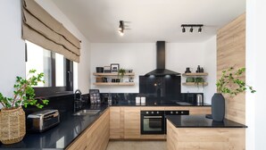 A modern kitchen with wooden cabinetry, a black countertop, a built-in oven, and a black range hood. There are wooden shelves with plants and kitchen items, and a large window with a beige valance. - Villa with pool and wellness for 8 persons, 4 bedrooms,  (Poreč)