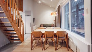 A modern interior with a wooden staircase, a dining area with a table and chairs, a sofa, and large windows with a view of trees. - Holiday Club Rukatunturi Apartments (Kuusamo)