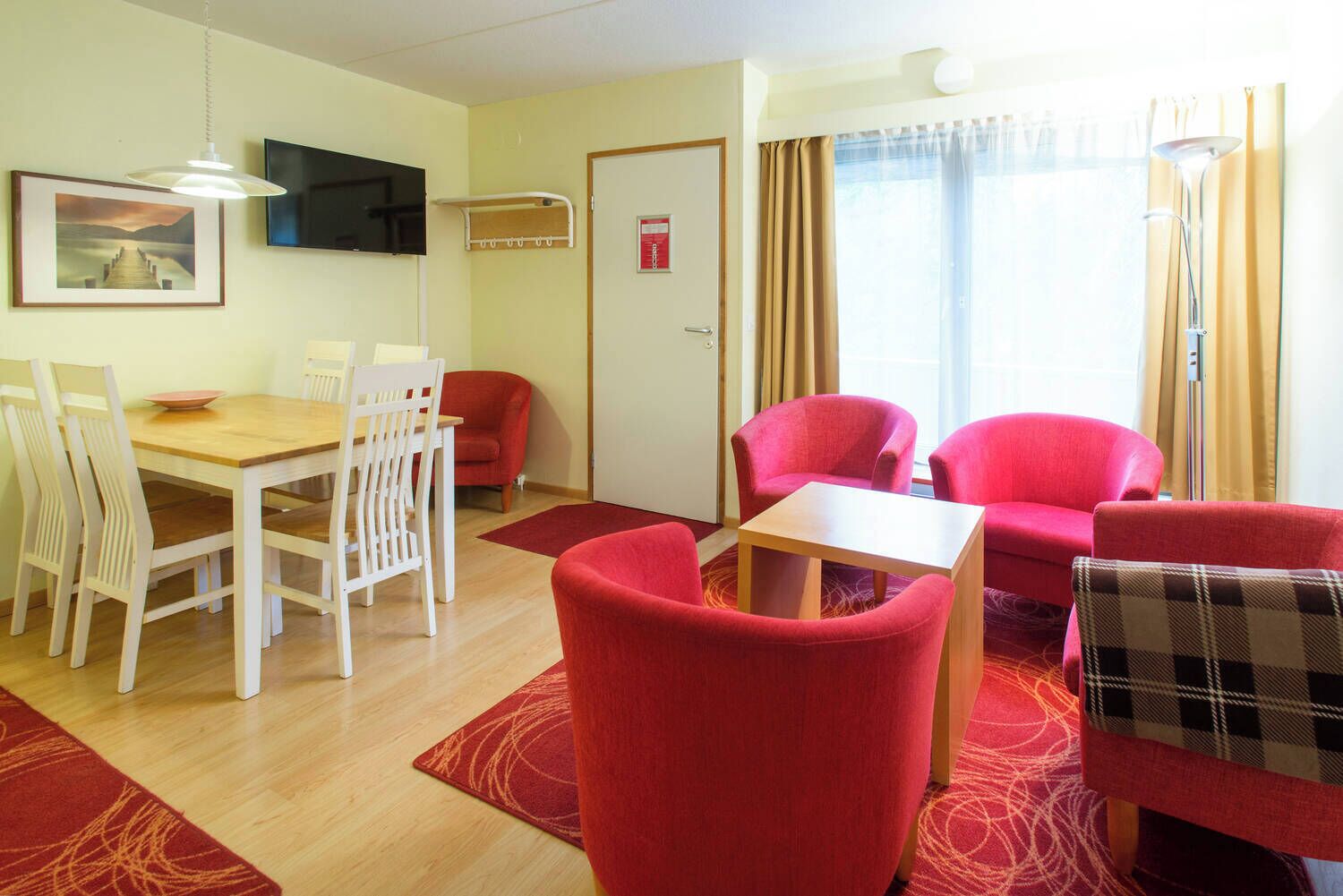A hotel room with a dining area, a TV mounted on the wall, a door with a fire safety sign, and a window with curtains.