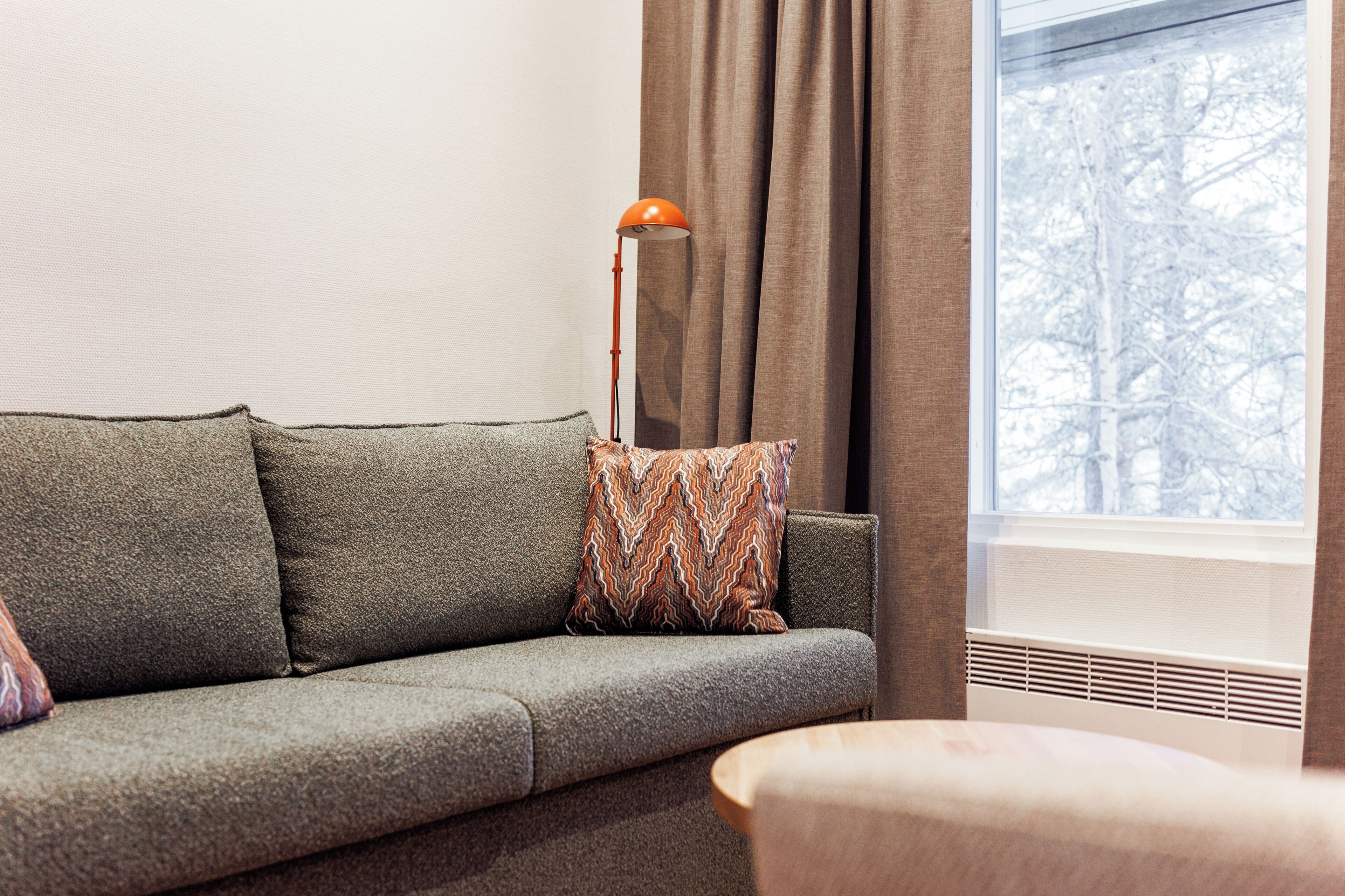 Un salon moderne avec un canapé gris, un coussin décoratif à motifs, une lampe sur pied et une vue sur un paysage enneigé par la fenêtre.