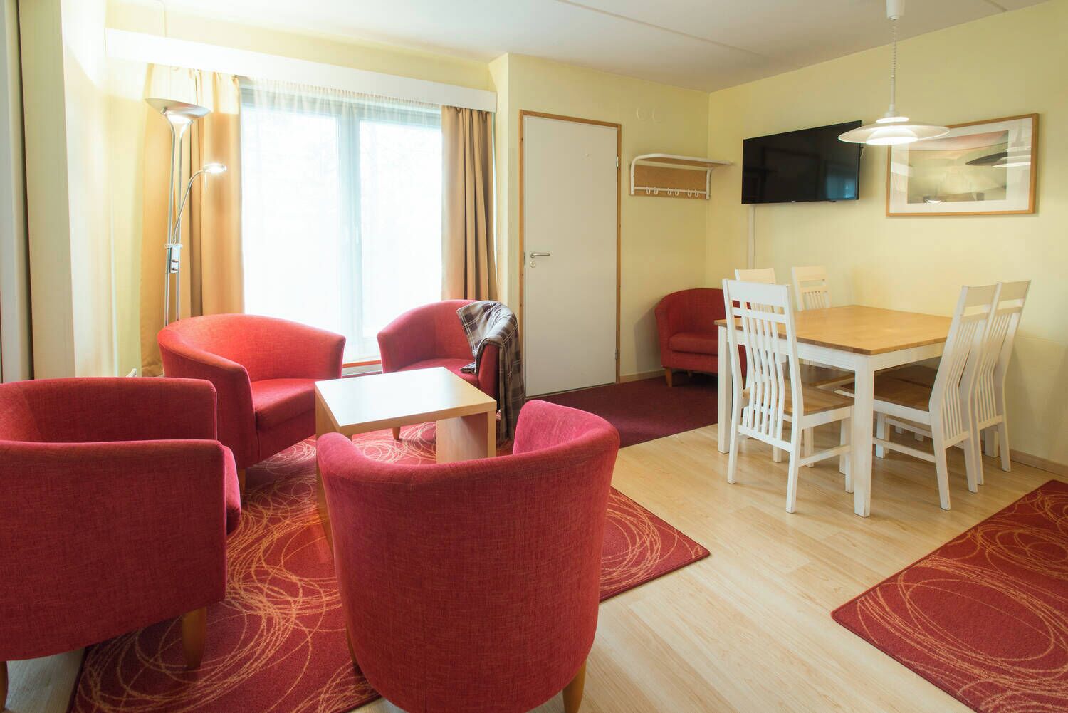 A hotel room with red upholstered chairs, a wooden table, a flat-screen TV, and a window with curtains.