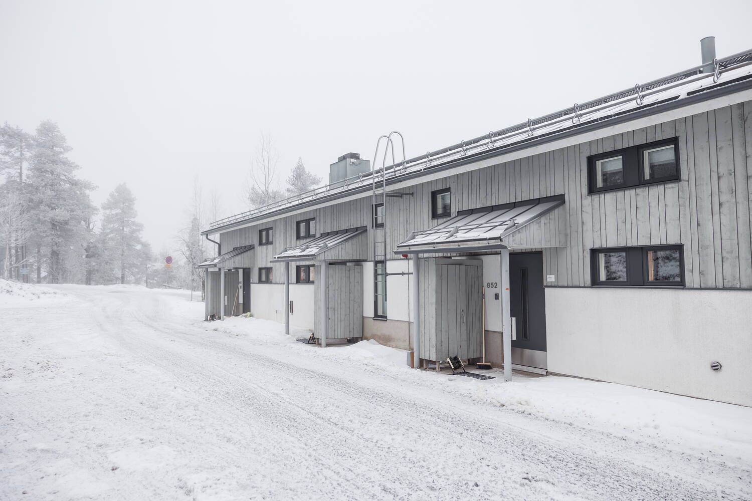 A row of wooden cabins with metal roofs, a snow-covered road, and trees in the background.