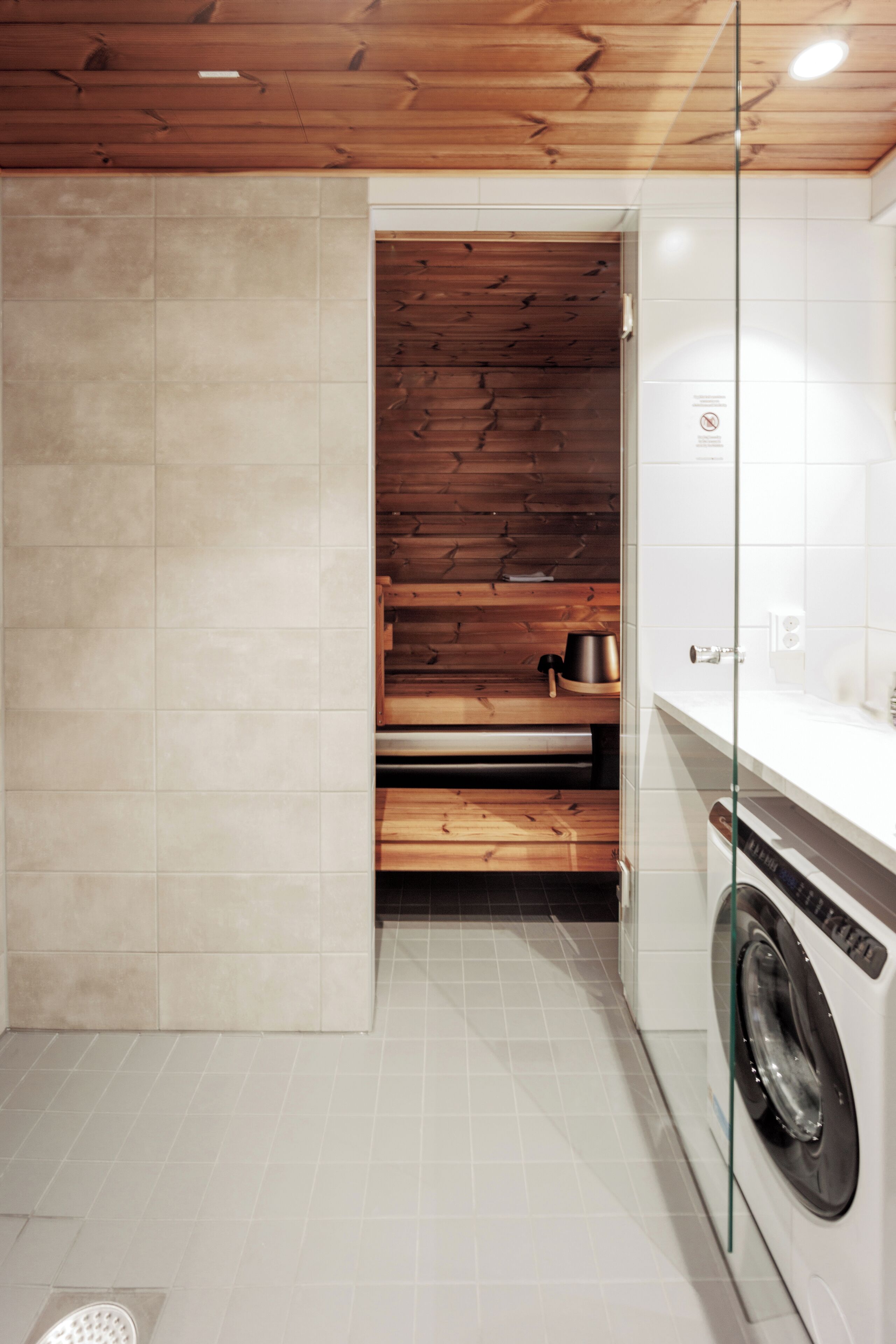 A compact bathroom with a wooden sauna, a washing machine, and a glass shower enclosure.
