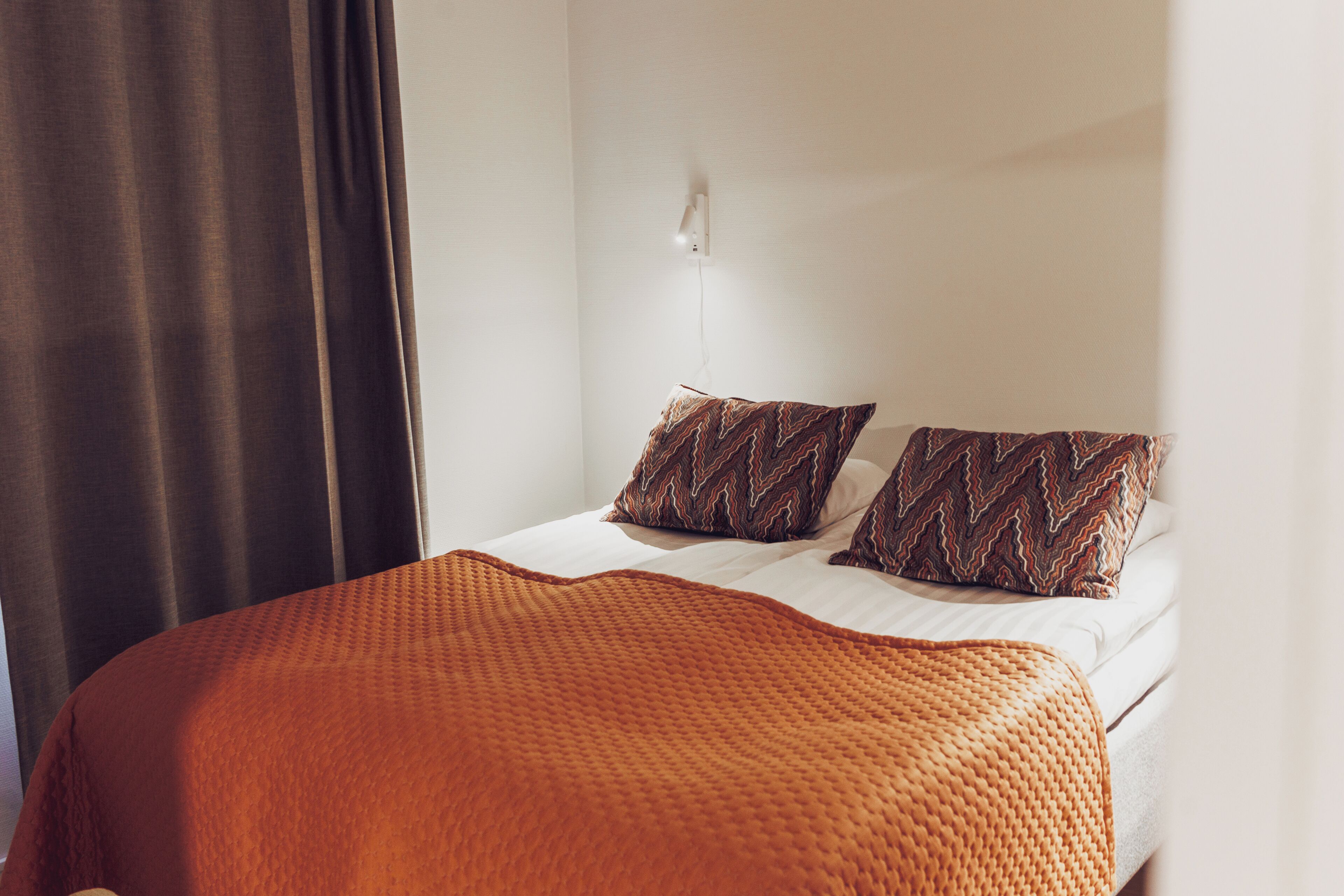 A neatly made bed with an orange blanket and two patterned pillows, a white wall, and a curtain.