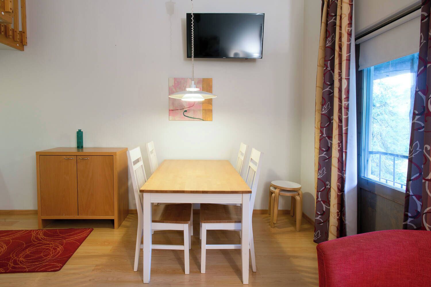 Un coin repas avec une table et des chaises en bois, un petit meuble, un téléviseur fixé au mur et une fenêtre avec des rideaux.