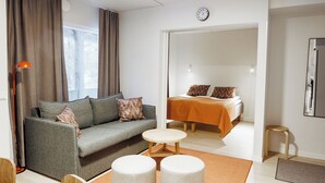 A modern hotel room with a sofa, a small round table, a bed with a headboard, a mirror, and a window with curtains. - Holiday Club Rukatunturi Apartments (Kuusamo)
