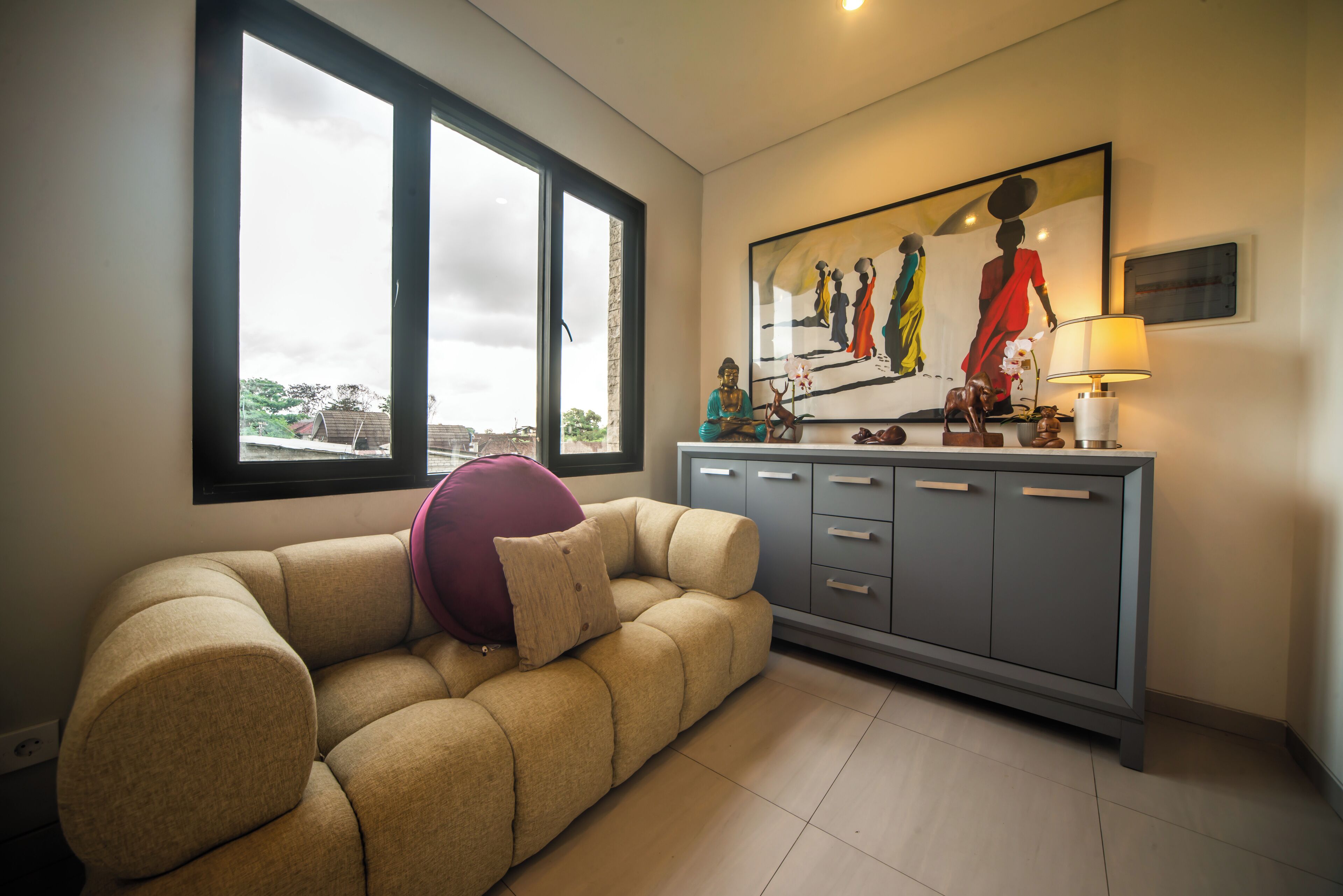 Een woonkamer met een beige bank, een paars kussen, een grijze kast en een schilderij met afbeeldingen van mensen aan de muur.