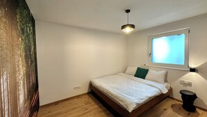 A bedroom with a bed, a window, a wall-mounted light, and a wooden floor.