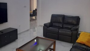 Ett vardagsrum med en svart lädersoffa, ett soffbord i glas, en tv-apparat placerad på ett svart skåp och en gul kudde.