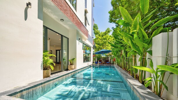 Una piscina con borde de baldosas, rodeada de plantas en macetas y un edificio blanco con grandes ventanales.