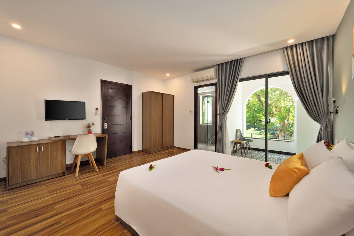 A hotel room with a bed, a desk, a chair, a TV, and a view of greenery outside.