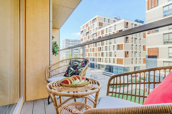 Un balcón con mobiliario de mimbre, una bandeja de fruta y vistas a edificios modernos.