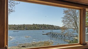 A wooden cabin with a large window overlooking a lake, a dock, and a forested shoreline. - Moon Tide Cottage | Oceanfront on Linekin Bay | Stunning Views (Boothbay Harbor)