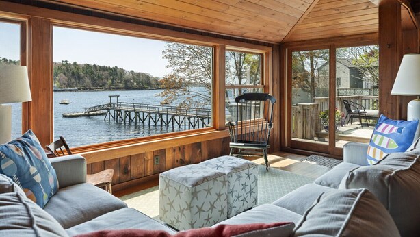 A cozy cabin with a wooden interior, large windows overlooking a lake and dock, and comfortable seating areas. - Moon Tide Cottage | Oceanfront on Linekin Bay | Stunning Views (Boothbay Harbor)