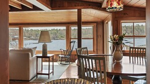 A cozy room with wooden walls and a large window overlooking a body of water. There is a table with chairs, a lamp, and a vase with flowers. - Moon Tide Cottage | Oceanfront on Linekin Bay | Stunning Views (Boothbay Harbor)