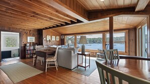A cozy cabin interior with a wooden ceiling, a fireplace, a sofa, a dining table, and a view of a lake. - Moon Tide Cottage | Oceanfront on Linekin Bay | Stunning Views (Boothbay Harbor)
