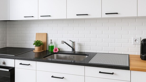 A modern kitchen with white cabinets, a black countertop, a stainless steel sink, a wooden cutting board, a potted plant, and a pink flower. - FriHome Centre Ville (Fribourg)