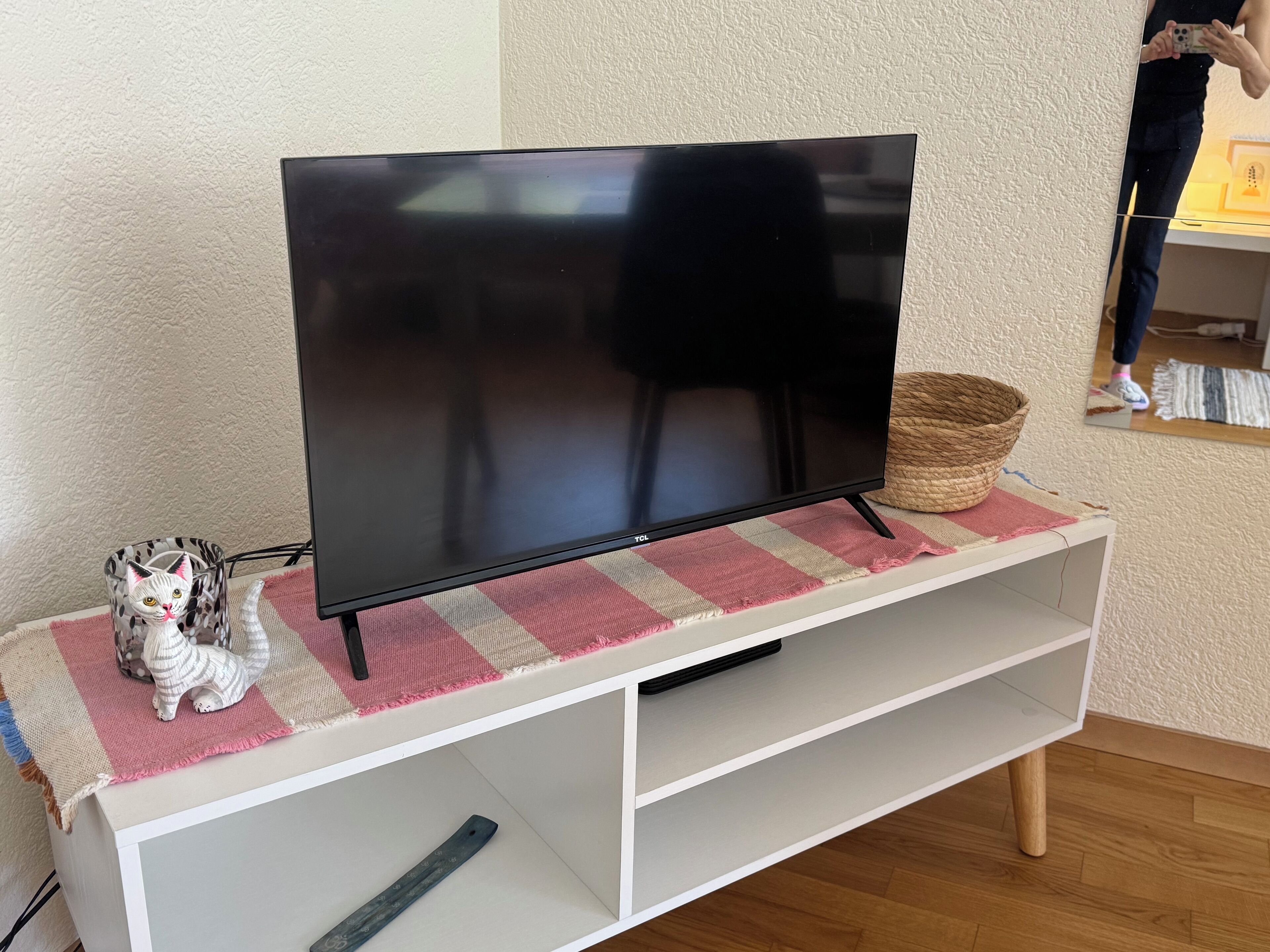 Litteänäyttöinen televisio valkoisella TV-jalustalla, jonka päällä on punottu kori ja raidallinen kangas.