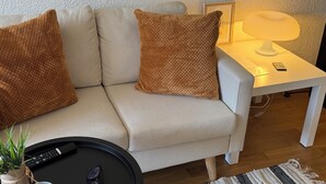 A beige sofa with two brown pillows, a small white side table with a lamp and remote control, and a black tray with a remote and sunglasses. - FriHome Centre Ville (Fribourg)