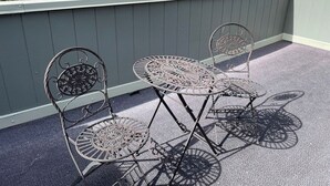 Un balcon avec deux chaises en métal et une table, une rambarde grise, et une vue sur les arbres et les bâtiments en arrière-plan.