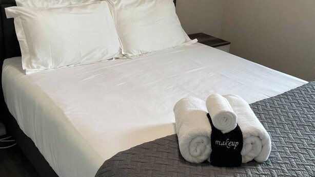 Un lit bien fait, avec des draps et des oreillers blancs, une couverture matelassée grise et deux serviettes enroulées, portant une étiquette noire indiquant « maquillage ».