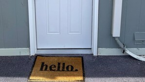 Une porte blanche portant le numéro 7, une petite fenêtre à carreaux et un paillasson sur lequel est écrit « bonjour ».