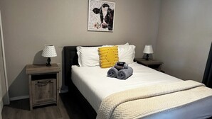 Un lit bien fait, recouvert d’une couette blanche, avec un oreiller jaune et des serviettes enroulées posées sur le côté. Un tableau encadré est accroché au mur, au-dessus du lit.
