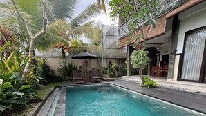 A rectangular swimming pool with a wooden deck surrounding it, set in a lush garden with various tropical plants and a building in the background. - Bayad Anyar Ubud Villa (Payangan)
