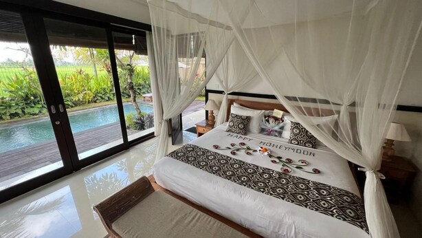A bedroom with a bed, a bedside table, a nightstand, and a view of a swimming pool. - Bayad Anyar Ubud Villa (Payangan)