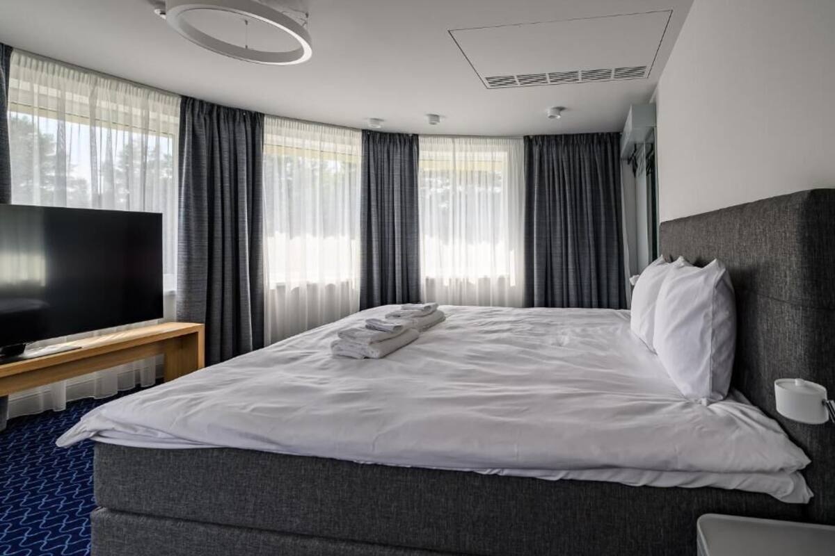 A hotel room with a large bed, a flat-screen TV on a wooden stand, a bedside table, and a window with curtains.
