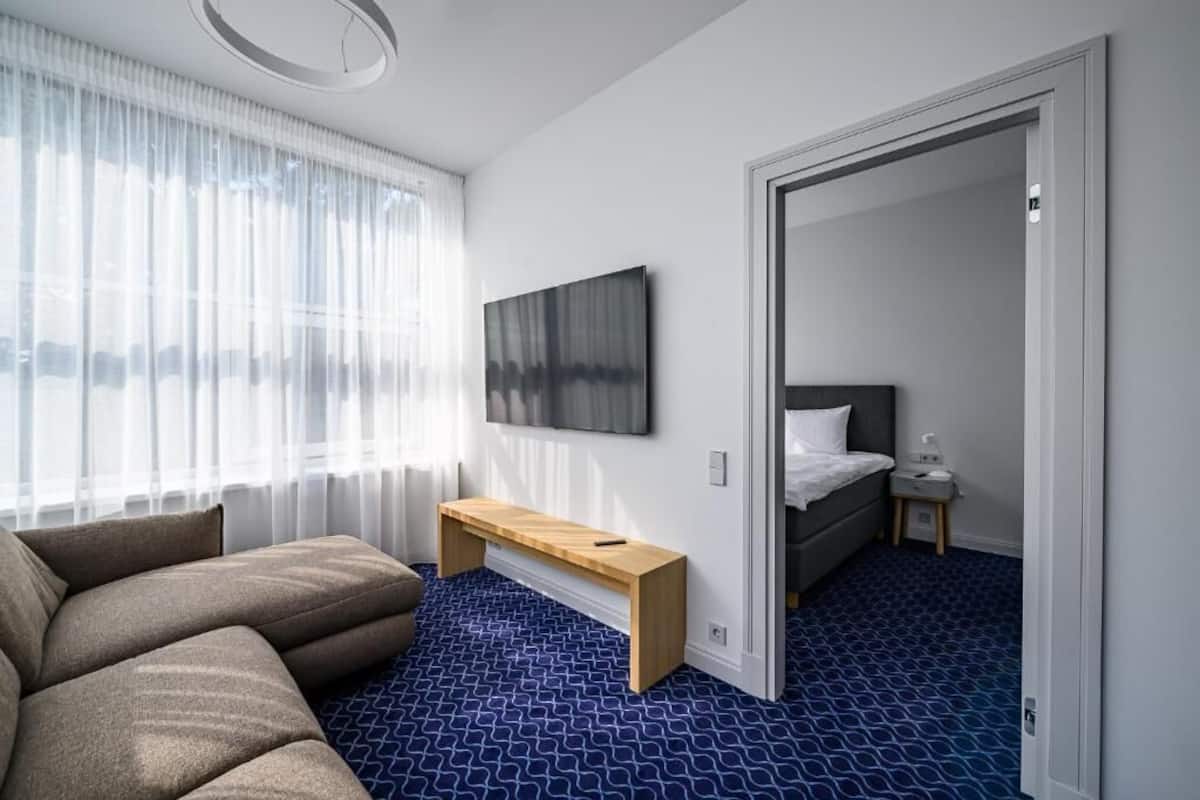 A modern hotel room with a sofa, a wooden bench, a bed, a bedside table, and a TV mounted on the wall.