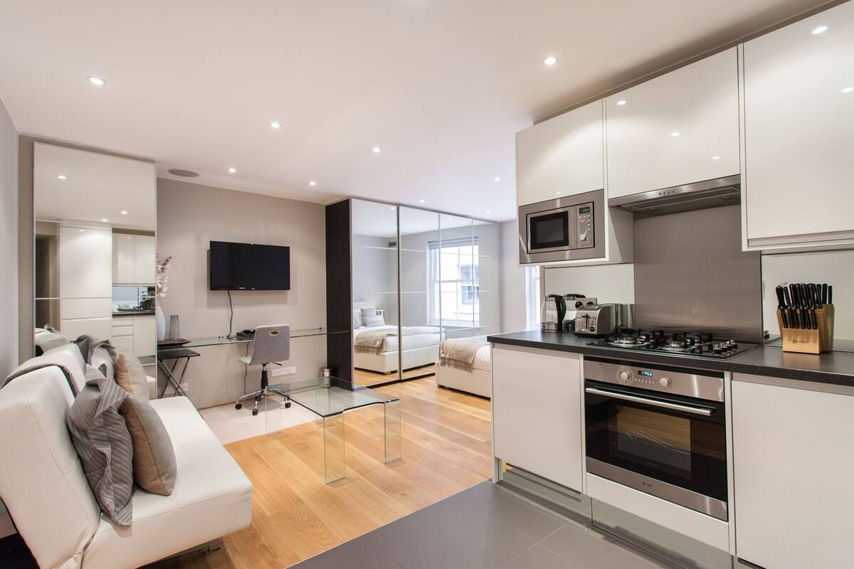 A modern kitchen with a built-in microwave, stove, and oven. There is a dining area with a glass table and chairs, and a sofa with a TV in the background.
