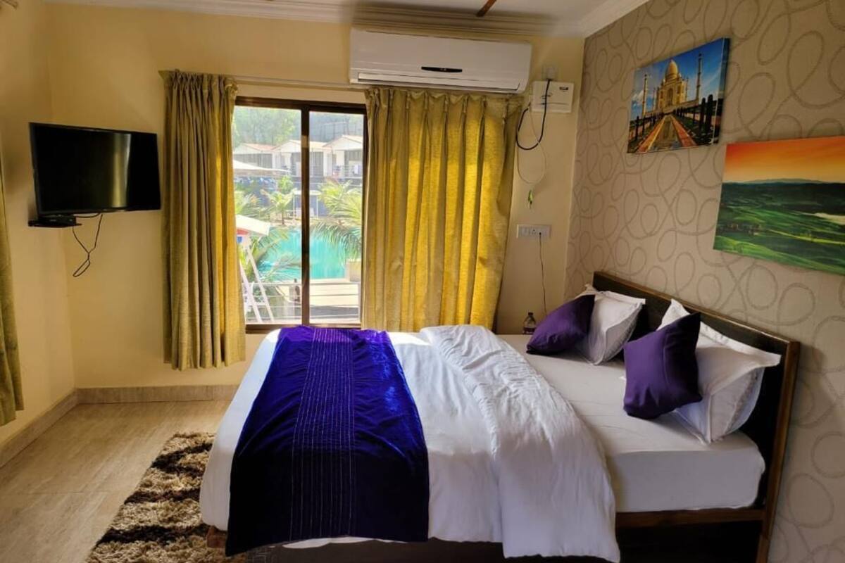 A hotel room with a bed, a TV, an air conditioning unit, a window with curtains, and a painting on the wall.