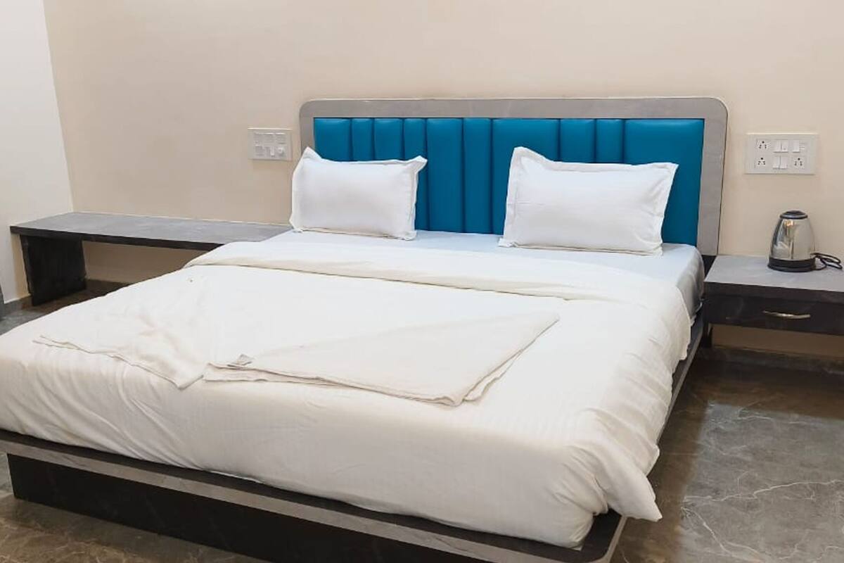 A neatly made bed with white bedding, a blue upholstered headboard, and a bedside table with a kettle.