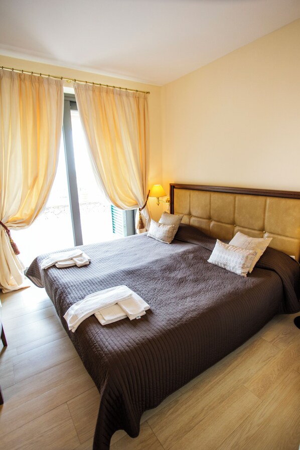 A neatly made bed with a brown headboard, a bedside lamp, and a window with curtains. - Mira-mare Apartments & Suites (La Spezia)