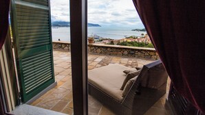 Un balcón con sofá de mimbre y cojines, con vistas a una ciudad costera, un muro de piedra y el mar.