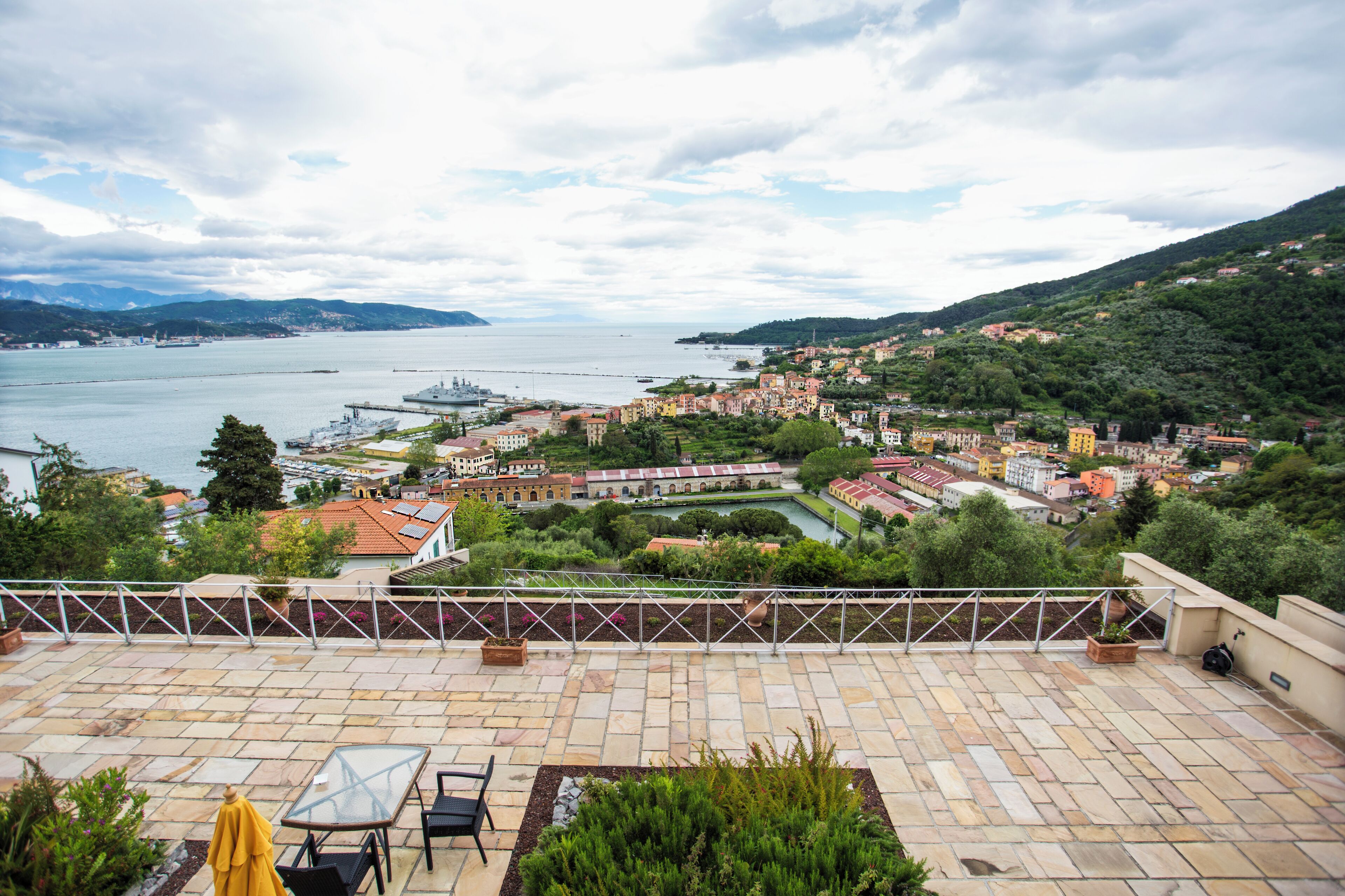 A terrace with a view of a coastal town, including buildings, greenery, and a body of water.