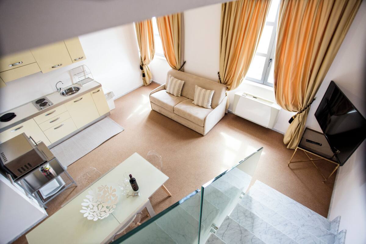 A modern kitchen with a built-in oven and microwave, a dining area with a glass table, a sofa, and a TV in a corner.